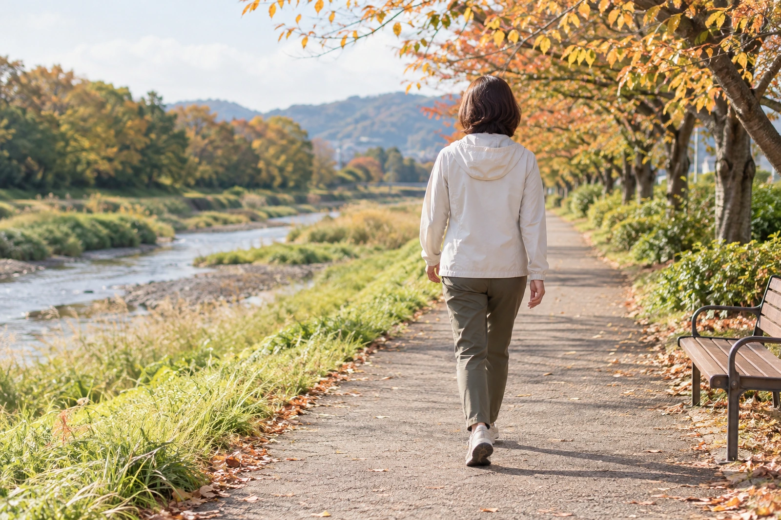 ベンチのある川沿いの道を一人で歩く50代女性の後ろ姿