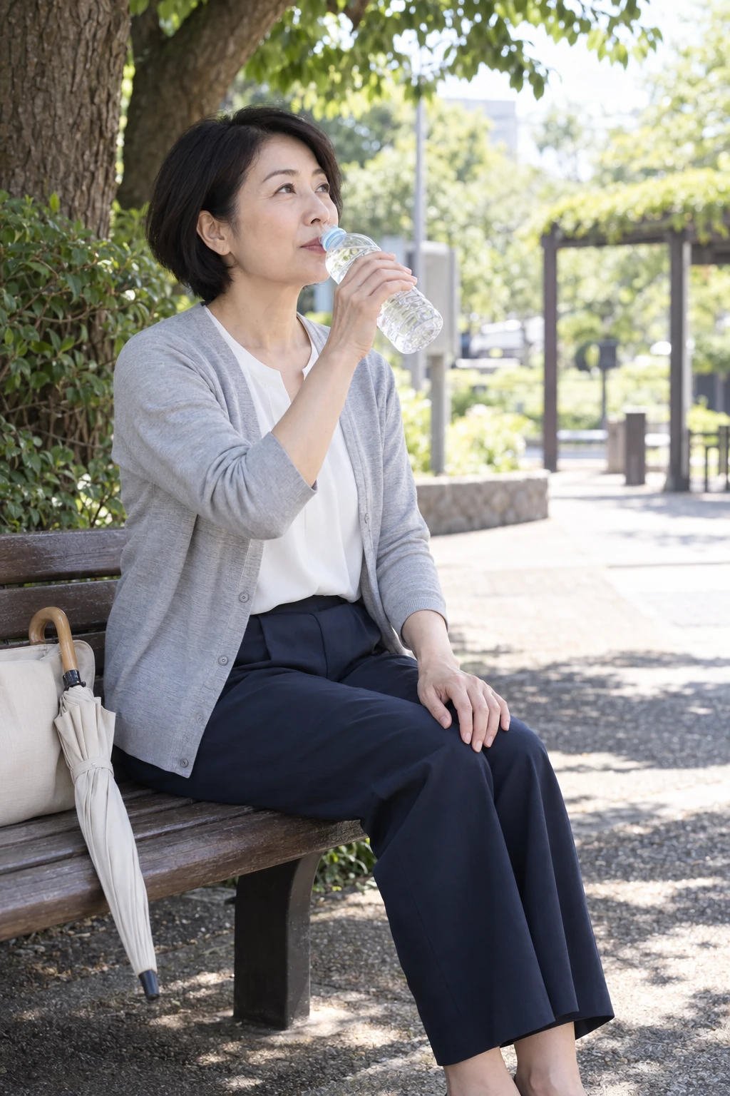 日陰のベンチで水分補給をして休む女性。閉じた日傘がそばに置かれている