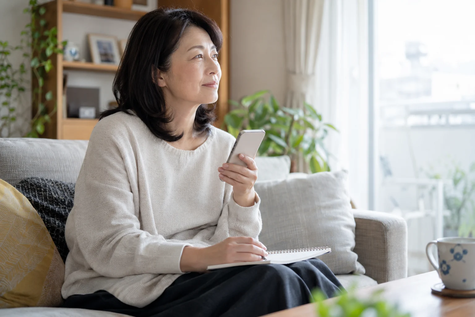 50代の日本人女性がスマートフォンとノートを持ち、会う前に尋ねたいことを整理している