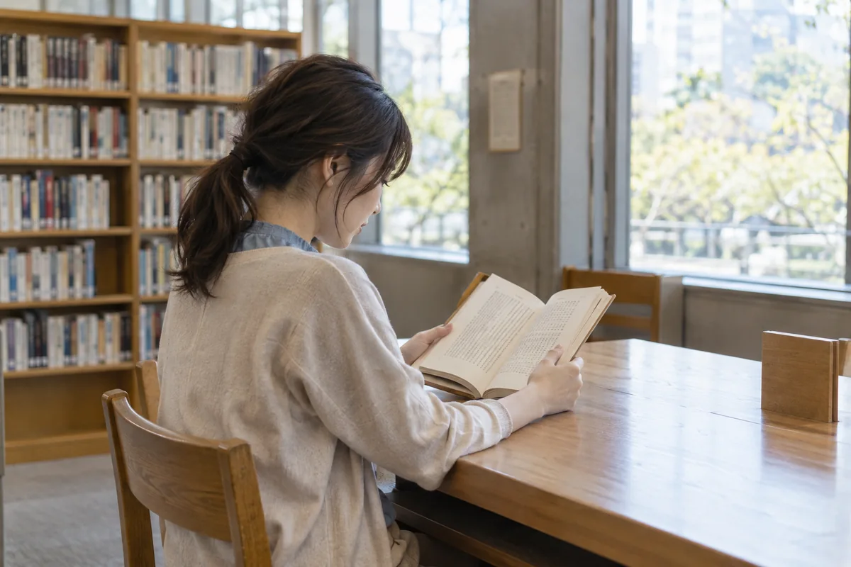 図書館でスマートフォンを見ずに一人で本を読む日本人女性の編集イメージ